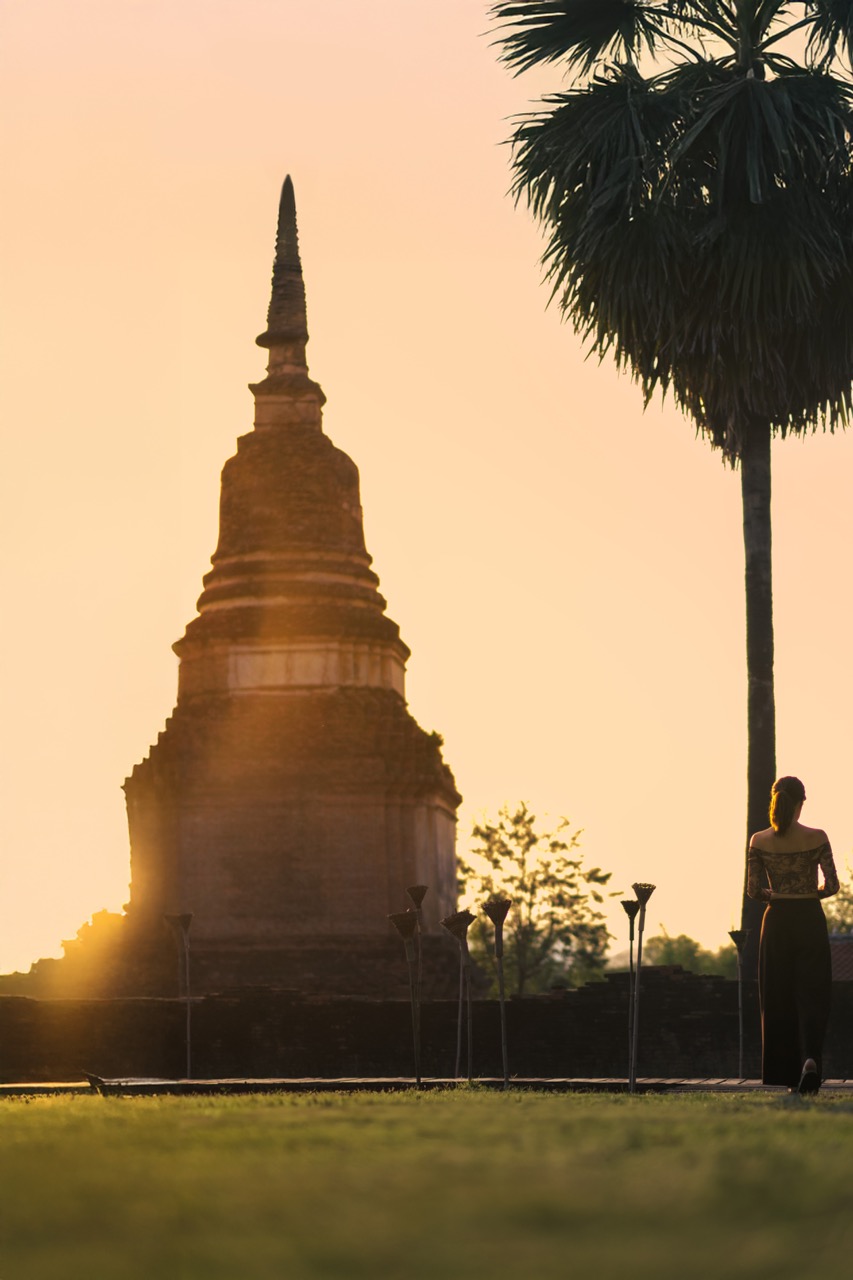 เจดีย์โบราณแห่งสุโขทัยเป็นเงาสะท้อนยามพระอาทิตย์ตก พร้อมคบไฟและต้นปาล์ม