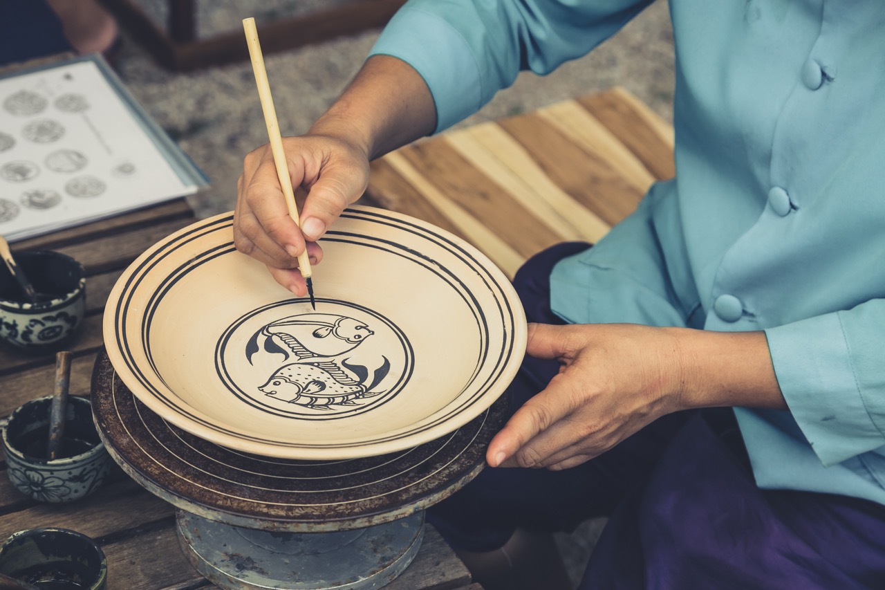 มือช่างฝีมือวาดลายปลากาดำแบบดั้งเดิมบนเครื่องปั้นดินเผาสังคโลกที่ยังไม่เคลือบ